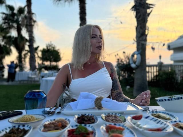 RedLara, con cabello rubio, vestido blanco y tatuajes, comiendo en una mesa al aire libre con una variedad de platos coloridos, con el atardecer de fondo, y un farol azul en la mesa..