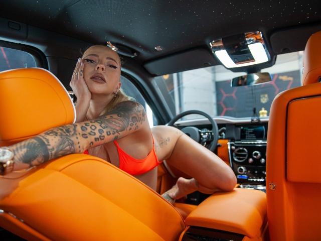 RedLara in een oranje bikini, leunend op gele autostoelen met een getatoeëerde arm, kijkend naar de camera, hand op haar wang, benen gespreid, haar achterwerk zichtbaar, in een moderne auto..