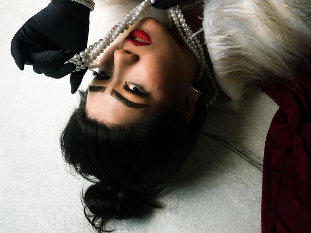 KhloeLawson with dark hair in a bun, wearing red lipstick, black gloves, pearl necklace, and fur, lying on white with her face upside down, looking at the camera.