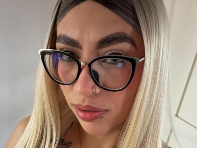 CamilleJuicyHot tiene cabello rubio, gafas negras, un piercing en la nariz y un piercing en el labio, y mira directamente a la cámara con los labios rosados ligeramente entreabiertos..