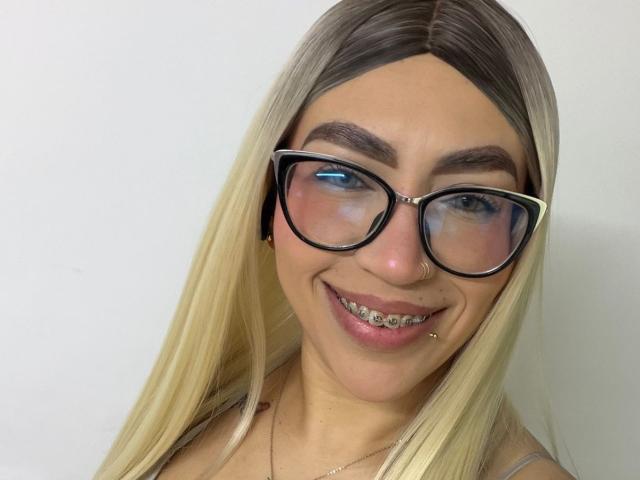 CamilleJuicyHot with blonde hair, glasses, nose ring, and a smile showing metal braces, wearing a thin necklace against a plain white background.