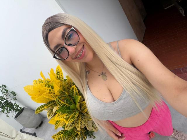Camille, com cabelo loiro, óculos, um top esportivo cinza e shorts rosa, exibindo seus seios grandes e sorrindo. Uma planta amarela em vaso ao fundo..