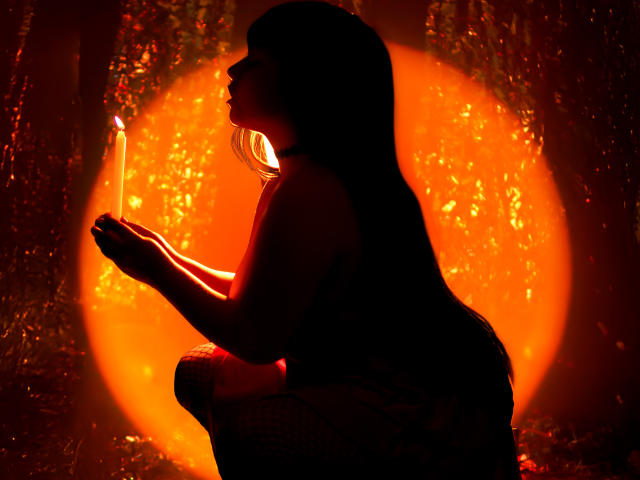 Silhouette de Nyxaria avec de longs cheveux noirs, agenouillée, tenant une bougie allumée, une lumière orange brillante derrière elle, les mains doucement jointes, une lumière douce sur le haut de son corps, une expression calme..