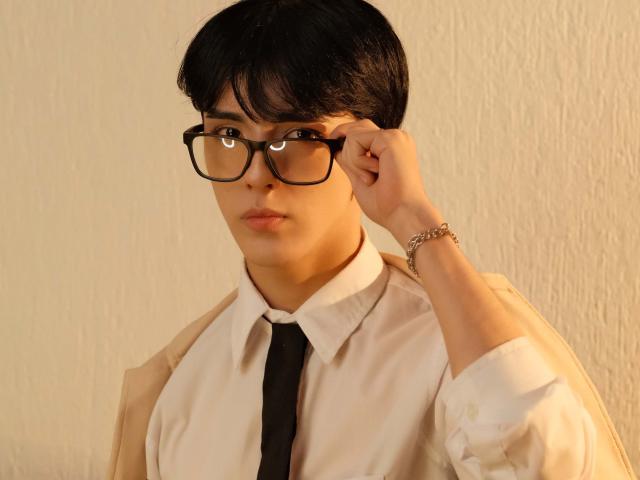 AerumChaee, con cabello negro corto, gafas y una expresión seria, vestida con una chaqueta beige sobre una camisa blanca con una corbata negra, ajustándose las gafas..