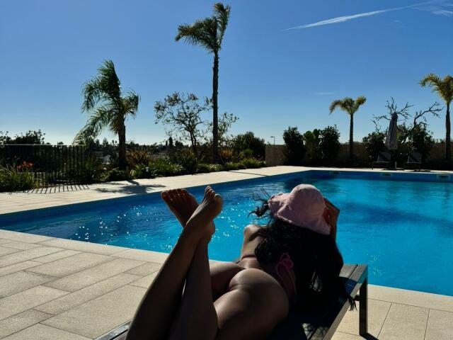 AmberMelisas is lying by a pool, legs up, wearing a pink hat, and a pink bikini, with her ass and bare feet in the clear blue water.