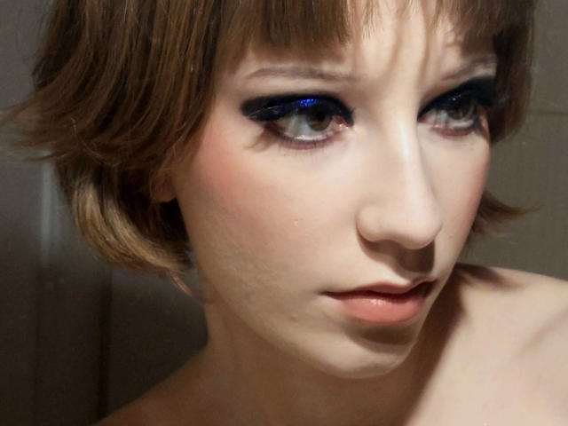 Alexa, sexy e attraente, con capelli castano corti, trucco occhi blu-nero, pelle chiara, labbra leggermente tumefatte e spalle scoperte, guarda verso il basso con un'espressione leggermente infastidita..