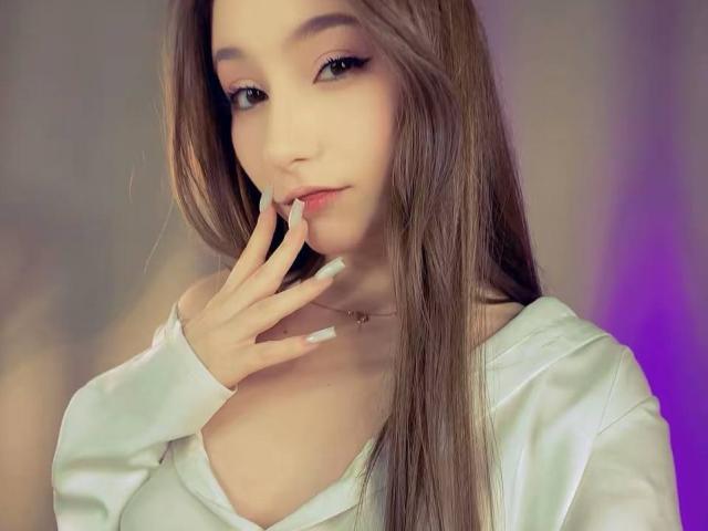 LunariaLive, con su largo cabello castaño, camisa blanca y piel pálida, tocándose los labios rosados con dedos con uñas blancas, mirando seductoramente a la cámara..