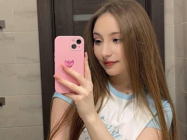 LunariaLive mit langen, braunen Haaren, hält ein rosa Handy mit Herz-Griff, trägt ein weißes und blaues Hemd, lächelt, macht ein Selfie im Badezimmerspiegel und zeigt ihr süßes Gesicht und ihre kleinen Brüste..