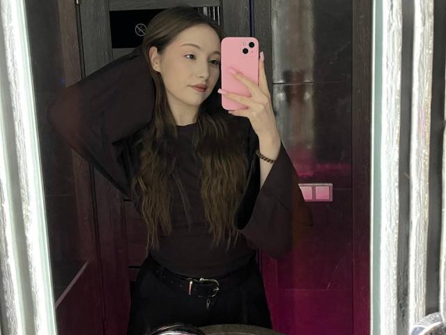 LunariaLive avec de longs cheveux bruns, un haut noir, un pantalon noir, un téléphone rose, bras levé, selfie devant un miroir, reflet de lumière rose, ceinture noire, maquillage discret, expression confiante..