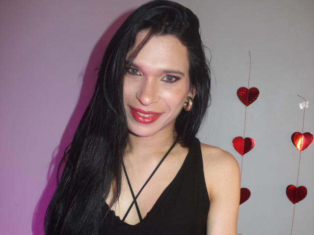 JadeOconner with long black hair, red lipstick, black sleeveless top, gold earrings, nose ring, smiling, white skin, pinkish-gray background, red heart decorations, and a string of small hearts.