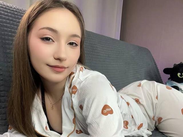 LunariaLive with light brown hair, pink eyeshadow, and white pajamas with brown hearts, lying on a gray couch, smiling softly, showing her cute face and small nose.