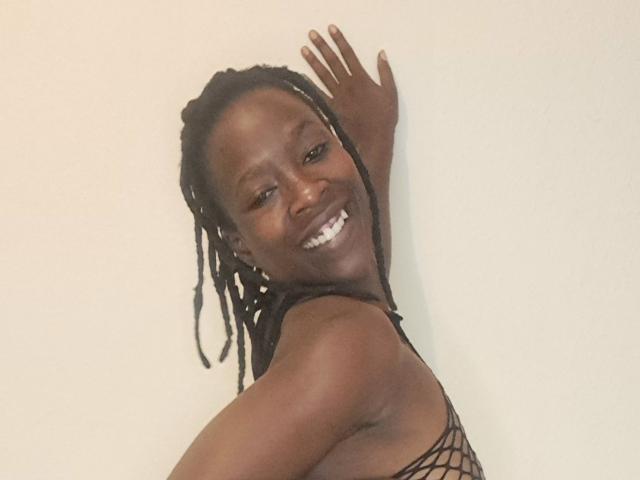 IrieItesEmpress, de piel oscura, sonriendo, mostrando los dientes, con un top de rejilla, rastas negras, brazo levantado, frente a una pared lisa, mirando por encima del hombro..