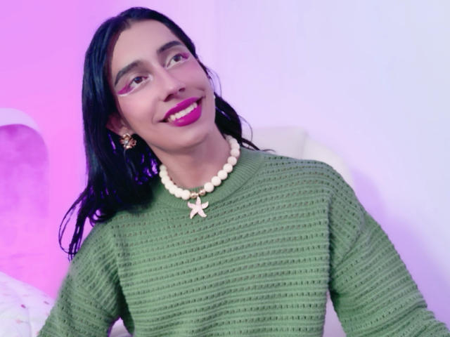 AlexaMissSx with black hair, green striped sweater, pink lipstick, white bead necklace, and pink star earring, smiling at camera. Soft pink and purple lighting.