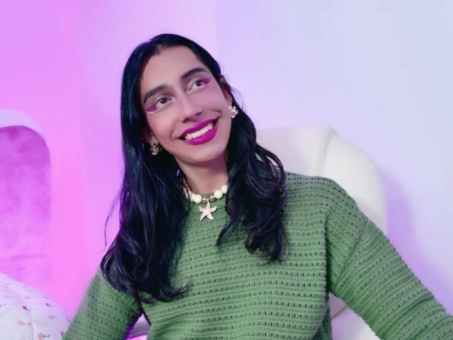 AlexaMissSx has long black hair, green knit sweater, pink lipstick, white chair, purple-pink lighting, and a star necklace, smiling with visible teeth.
