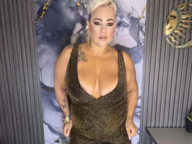 GianellaCox with platinum blonde hair, large tits in a deep green, glittery dress, tattoos on shoulders, colorful bracelet, standing, looking at camera, gray vertical background.