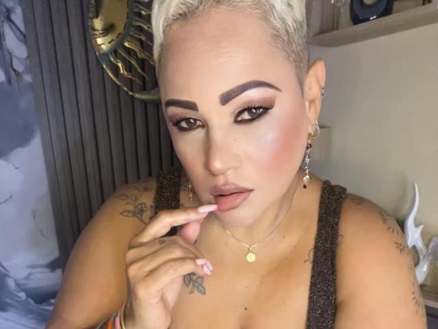 GianellaCox with bleached blonde hair, thick eyebrows, and full makeup, sucking her finger, showing her tits, and tattoos on her shoulder. She's wearing a gold necklace and earrings.