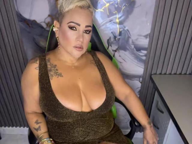 GianellaCox with large tits in a sparkly brown dress, sitting on a green chair, blonde pixie cut, tattoos on shoulder and arm, wearing gold earrings and bracelets.