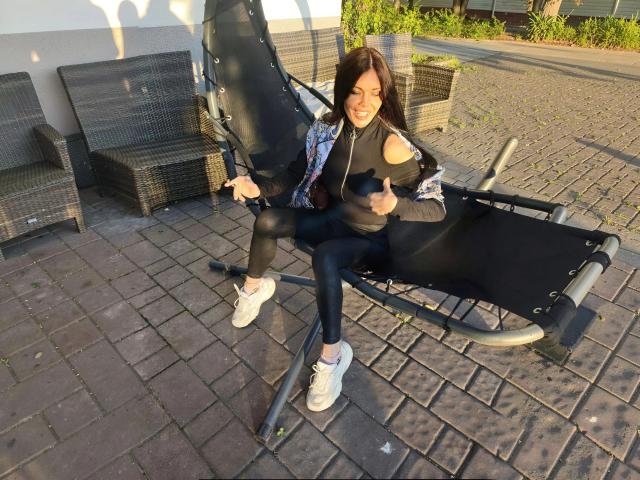 MissLover assise sur un fauteuil de relaxation en métal noir, faisant deux pouces levés, portant une veste noire avec des épaules découpées, des leggings noirs et des baskets blanches, sur une terrasse en briques..