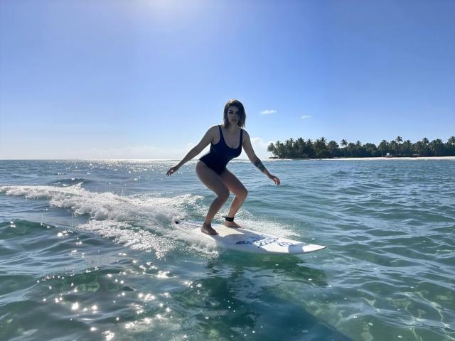 MissLover surft auf einem weißen Surfbrett, trägt einen schwarzen Badeanzug, hat einen tätowierten Arm, und im Hintergrund erstreckt sich ein sonnendurchfluteter, klarer, blauer Ozean. Ihre Brüste sind leicht unter dem Wasser sichtbar..