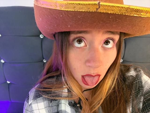 DannaGomezLove with wide eyes, sticking out red lipstick-covered tongue, wearing a brown, glittery sombrero, and a black-and-white checkered shirt. Her brown hair is straight.