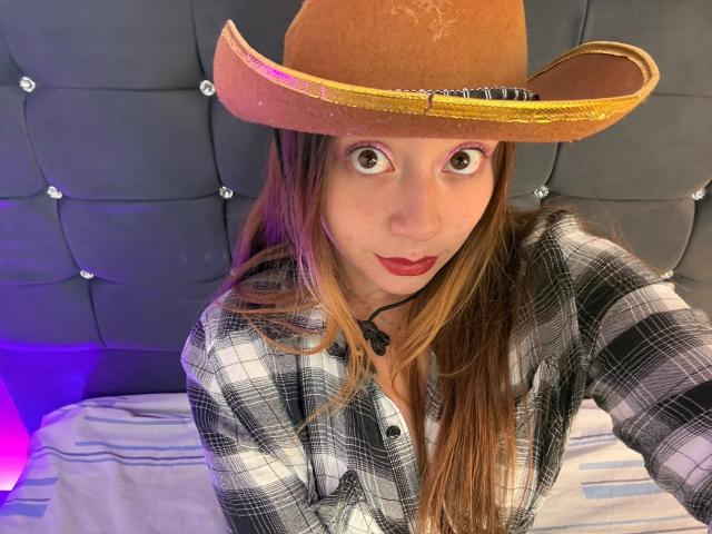 DannaGomezLove with long brown hair, wearing a brown cowboy hat with yellow trim, red lipstick, and a black-and-white plaid shirt, sitting on a bed.