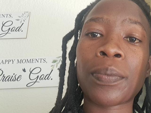 IrieItesEmpress hat eine dunkelbraune Haut, schwarze Dreadlocks und lächelt mit leicht geöffneten Lippen, wobei ein wenig von ihren Zähnen zu sehen ist. Sie schaut direkt in die Kamera..