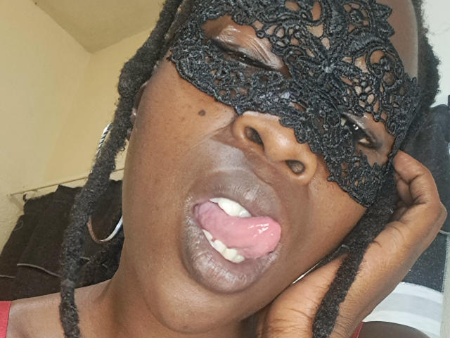 IrieItesEmpress with dark skin, wearing a black lace mask, tongue out, and red lips, has dreadlocks and a small mole near her right eye.