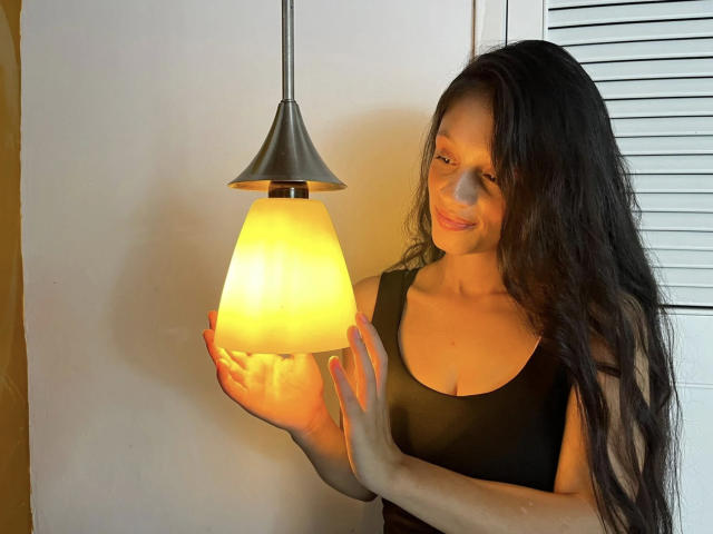 CloeLovely con lunghi capelli neri, che tiene una lampada gialla luminosa, indossa una canotta nera, sorride dolcemente, con le mani sulla lampada, uno sfondo con un muro bianco e delle persiane..