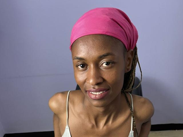 CandyCross, een donkerhuidige vrouw met een roze hoofddoek, kleine borsten, die lacht, gekleed in een witte tanktop, met vlechten, zittend op een zwarte stoel, met een lichtpaarse achtergrond..