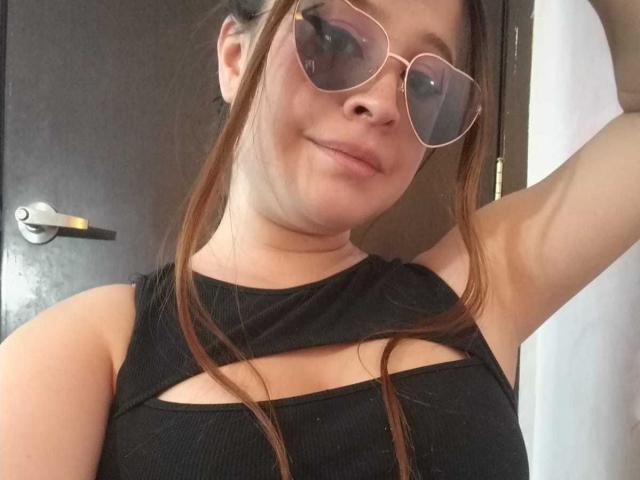 Melani Becker, avec ses cheveux bruns, portant de grandes lunettes roses, un haut noir découpé qui révèle ses seins, souriante, bras levé, debout devant une porte sombre..
