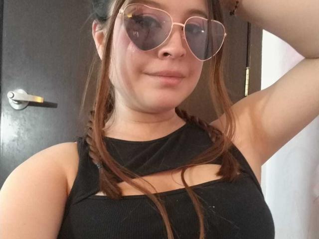 MelaniBecker with brown braids, wearing black sleeveless top with cutout, and gold aviator sunglasses, smiling slightly, raising arm, showing her tits and a small nose bruise.
