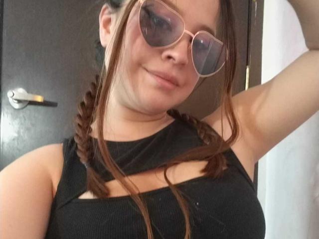MelaniBecker with braids, wearing a black top, has tits and is smiling at the camera, wearing pink sunglasses, arm raised.