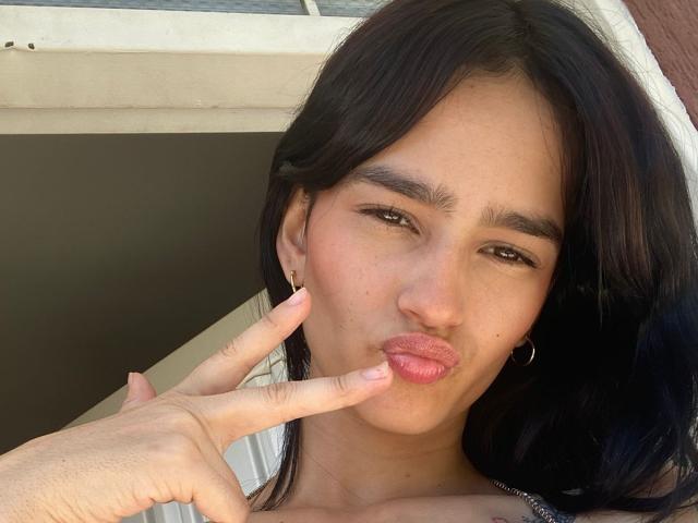 MelodyMansson with dark hair, brown eyes, and pink lips making a kissy face, touching her lips with her finger. She's wearing hoop earrings and has a slight tan.