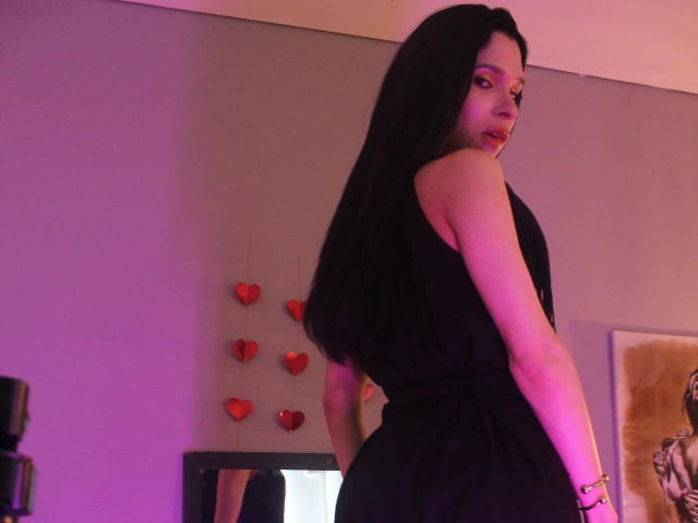 JadeOconner with long black hair, wearing a black dress, looking over her shoulder, purple and pink lighting, red heart wall decorations, bracelet on right wrist, standing in room.