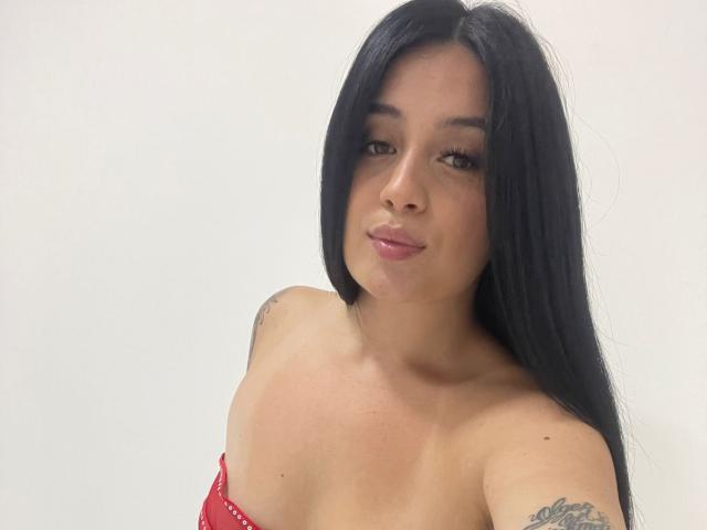 AnnyBentleii, con cabello negro largo, ojos marrones y lápiz labial rosa, vestida con un top rojo sin tirantes, mostrando tatuajes en sus brazos, sonriendo ligeramente y dejando ver sus pequeños senos..