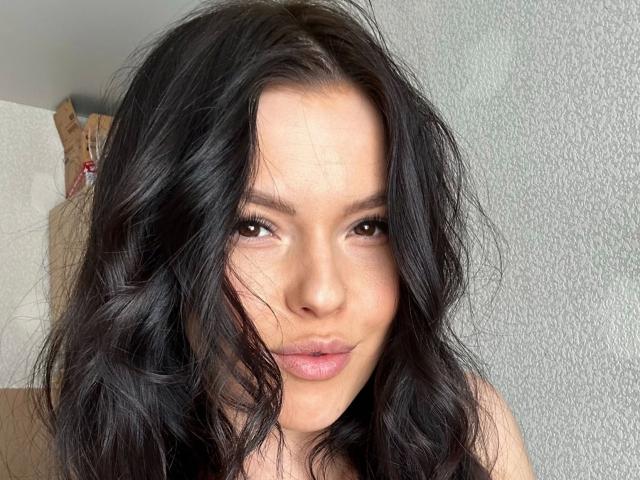 Eva24-hot ha i capelli scuri e ondulati, gli occhi marroni e le labbra piene di colore rosa. Il suo viso è centrato e guarda leggermente di lato, con un'espressione appena accennata. La sua pelle è chiara..