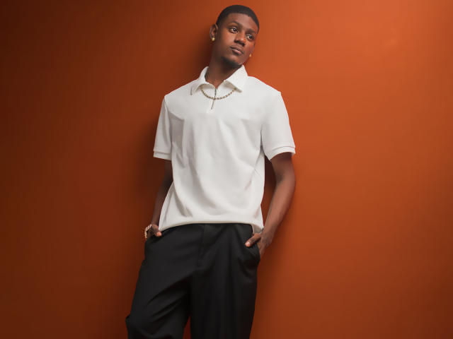 DrakeWit, black dude, white polo, black pants, hands in pockets, leaning against orange wall, short curly hair, earrings, serious expression, slim build, subtle smile, relaxed pose.