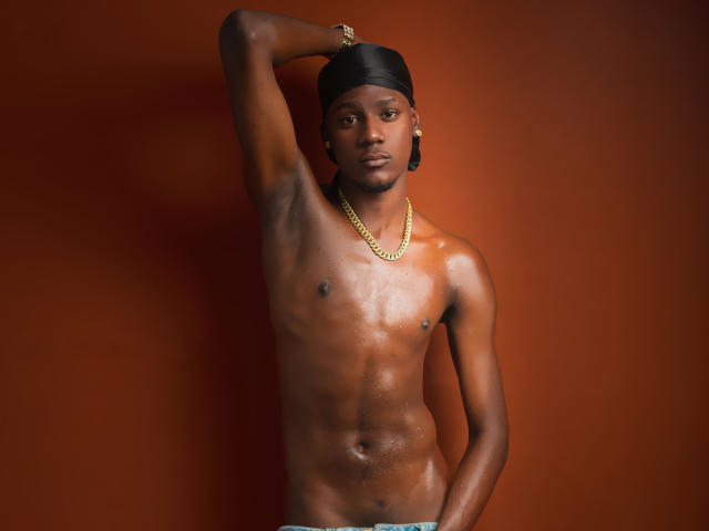 Glänzender, verschwitzter DrakeWit mit einem schwarzen Kopftuch, einer Goldkette und ohne Kleidung, steht vor einer in verbranntes Orange gehaltenen Wand, ein Arm erhoben, die Hand auf dem Kopf, und blickt in die Kamera..