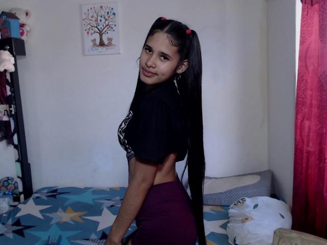 ScarlettRoss69 met lange, zwarte vlechtjes, gekleed in een zwarte top en paarse broek, kijkt om en heeft een schattige glimlach, waarbij haar kleine billen en slanke taille zichtbaar zijn in een slaapkamer..