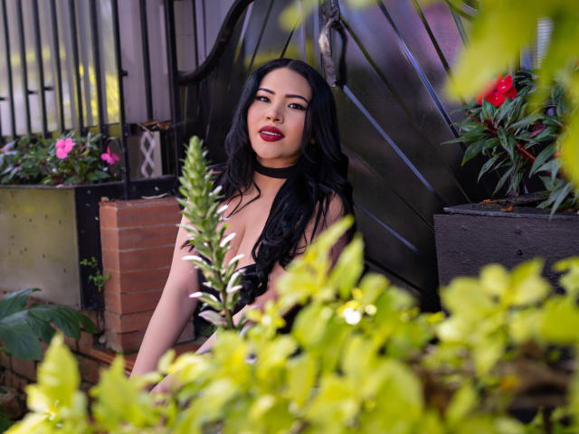 OliviGrace, avec ses longs cheveux noirs, son rouge à lèvres rouge et son collier noir, assise dans un jardin verdoyant, montrant ses seins, apparaissant sexy et confiante..