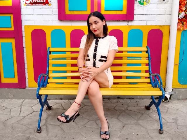 MindyLove sitting on a yellow bench with blue metal legs, wearing a white dress with black buttons, black sandals, and a hair clip, with her legs crossed, smiling.