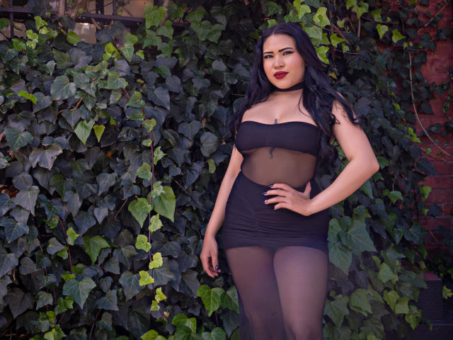 OliviGrace with black hair, red lipstick, and sheer black dress showing her tits and pussy, standing in front of leafy green vines. She has a hand on her hip.