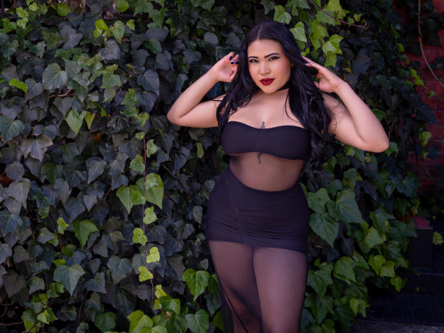 OliviGrace with dark hair, wearing a black strapless top and sheer black skirt, standing against green ivy, hands in hair, red lipstick, and a confident, sexy expression.