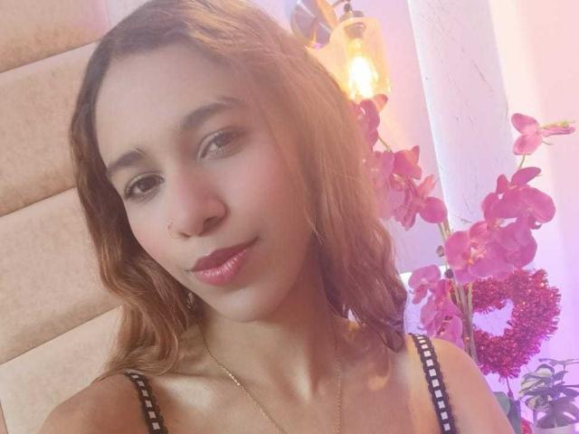 Valerie Woods, con i capelli castano ondulati, la pelle chiara e il rossetto rosa, indossa una maglietta a righe nere e bianche, mostrando il suo nasino piccolo e il suo sorriso delicato, vicino a orchidee rosa..