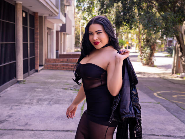 OliviGrace with long black hair, red lipstick, and a strapless black sheer dress, holding a black puffy jacket, standing on a sunlit sidewalk with trees in the background.