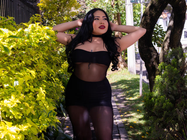 OliviGrace, avec ses longs cheveux noirs, son rouge à lèvres rouge foncé, portant un haut noir transparent et une jupe noire moulante, debout, les mains dans les cheveux, sexy, pulpeuse, et montrant ses fesses..