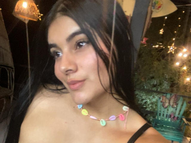 AlaraKey with dark hair, light skin, and a nose ring, wearing a colorful shell necklace, black strap dress, and subtle makeup, smiling slightly. Boobs partially visible.