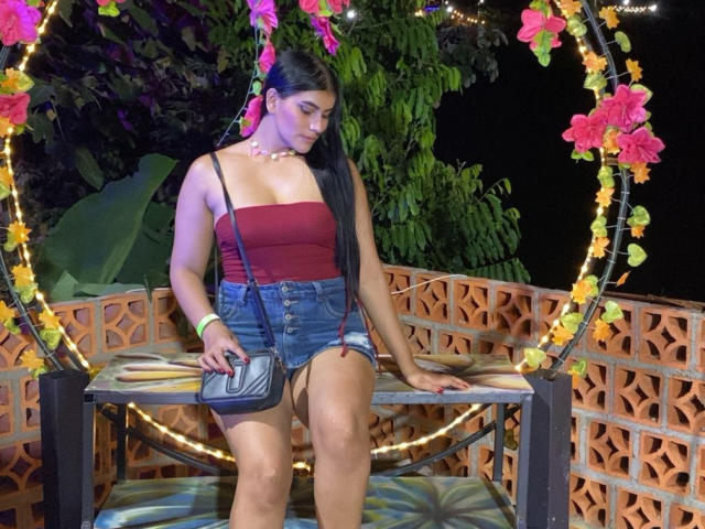 AlaraKey with long black hair, red tube top, and denim shorts, sitting on a decorated swing, holding a black purse, showing her tits and ass, wearing a white necklace.