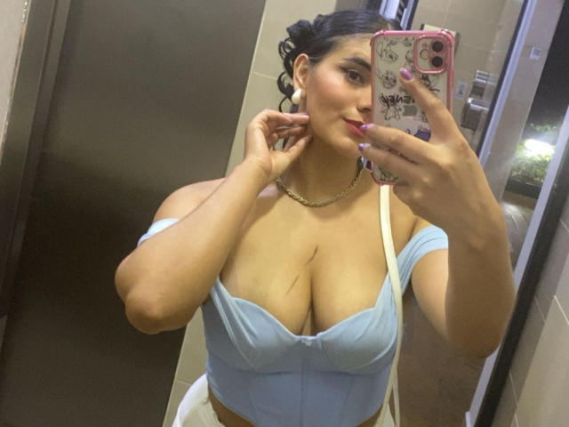 AlaraKey with big tits in a light blue off-shoulder top, holding a pink phone, taking a mirror selfie, showing her cleavage, wearing a chain necklace, in a bathroom.