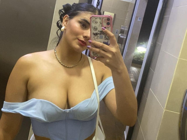 AlaraKey with large tits, wearing a blue off-shoulder top, holding a phone with a pink case, in a bathroom, has a gold chain, and white earrings.